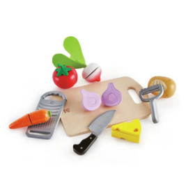 Spiel-Schneidebrett mit Spielgemüse, Käse, Messer, Reibe und Schäler auf weißem Hintergrund arrangiert.