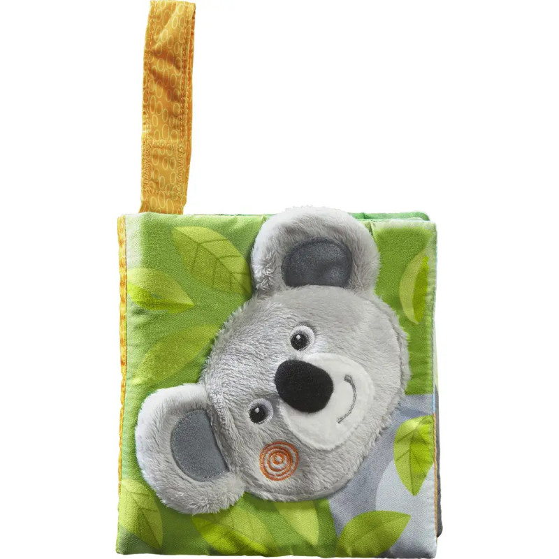 Weiches Babybuch mit Plüsch-Koala-Gesicht und gelber Stoffschlaufe vor grünem, blättrigem Hintergrund. Weiches Babybuch mit Plüsch-Koala-Gesicht und gelber Stoffschlaufe vor grünem, blättrigem Hintergrund.