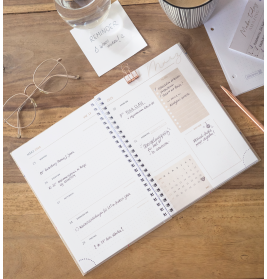 Offener Planer auf Holztisch mit Brille, Kaffee, Notizen und einem Glas Wasser mit Erinnerungsetikett.