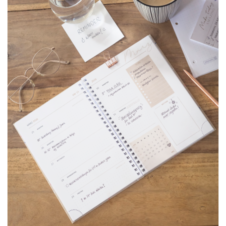 Offener Planer auf Holztisch mit Brille, Kaffee, Notizen und einem Glas Wasser mit Erinnerungsetikett.