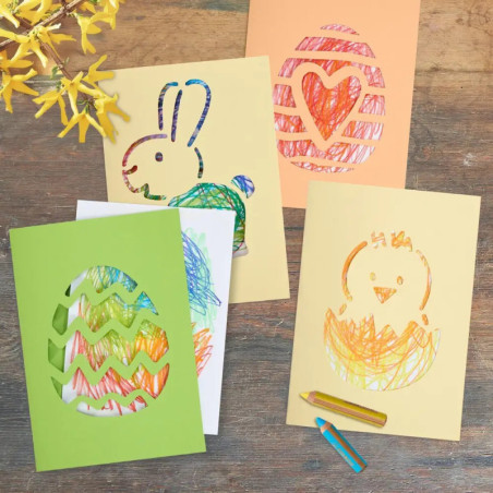 Vier bunte Ostkarten mit Wachsmalbildern von Ei, Hase, Herz und Küken liegen auf einem Holztisch.