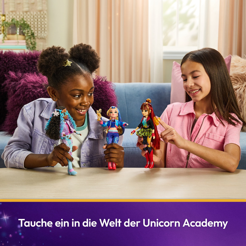 Zwei Mädchen spielen lächelnd am Tisch mit Unicorn Academy Puppen, vor einem lila Hintergrund.