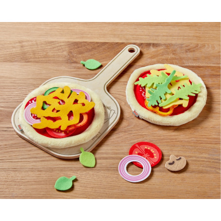 Zwei Filzpizza-Spielzeuge mit bunten Belägen auf Holzbrett und Tisch, umgeben von einzelnen Toppings.
