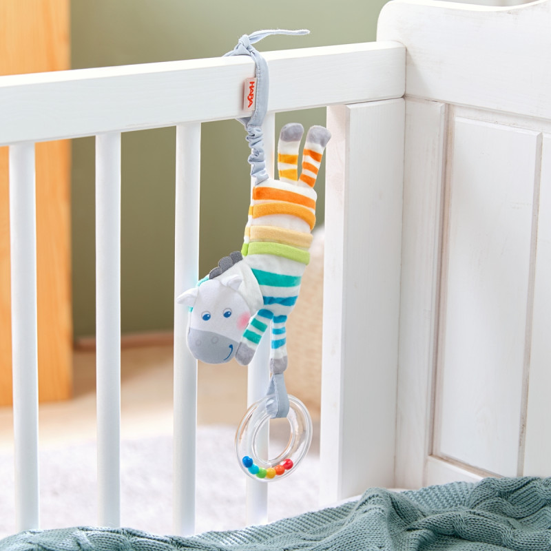 Plüschzebra mit bunten Streifen hängt am weißen Gitterbett und hält einen durchsichtigen Rasselring.