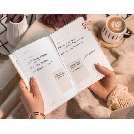Frau hält offene Planer mit Notizen, daneben steht eine Tasse Kaffee mit Latte Art.