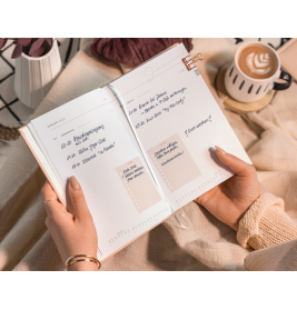 Person mit geöffnetem Planer mit handschriftlichen Notizen neben einer Tasse Kaffee mit Latte Art.