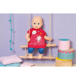 Eine Babypuppe im roten Kleid sitzt auf einer Holzschaukel, daneben liegen Kissen und bunte Kisten.