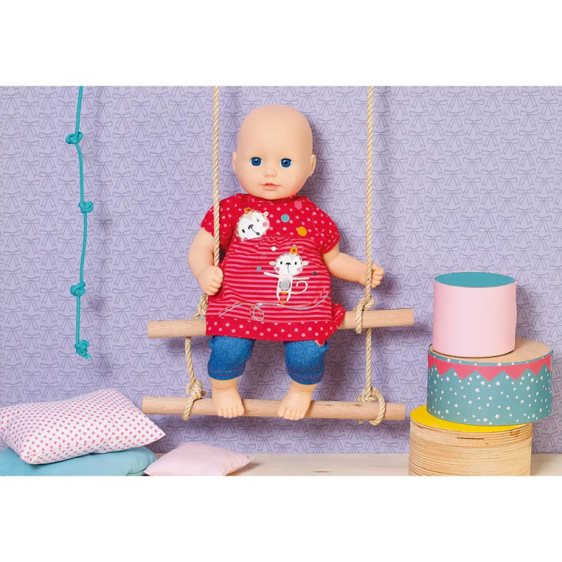 Eine Babypuppe im roten Kleid sitzt auf einer Holzschaukel, daneben liegen Kissen und bunte Kisten.