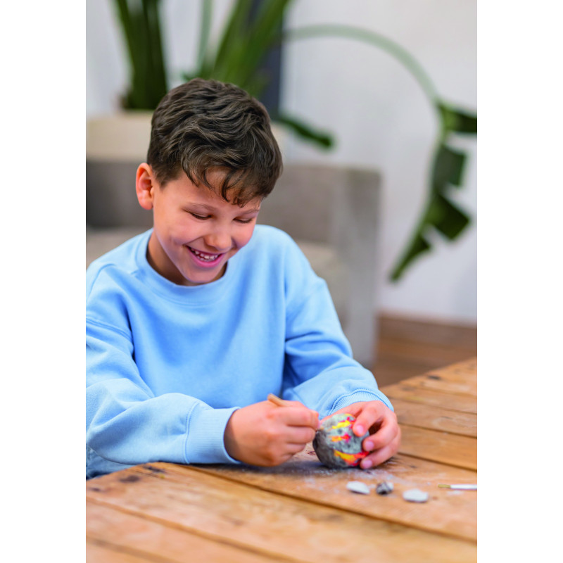 Lächelndes Kind im blauen Sweatshirt bearbeitet mit einem Werkzeug einen bunten Stein an einem Holztisch.