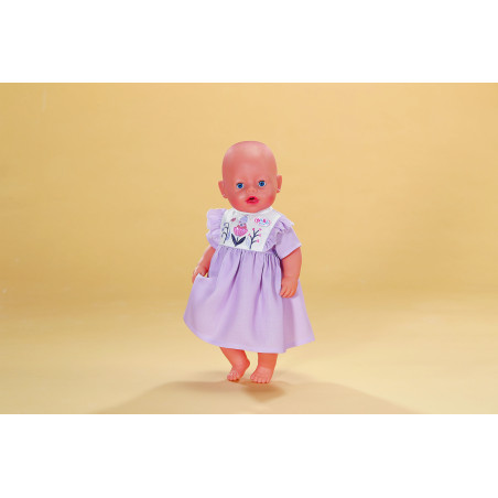 Eine glatzköpfige Babypuppe im lavendelfarbenen Kleid steht barfuß vor beigem Hintergrund.