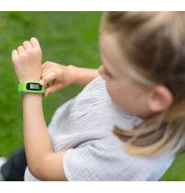 Ein Kind betrachtet im Freien auf dem Rasen eine grüne Digitaluhr an seinem Handgelenk.