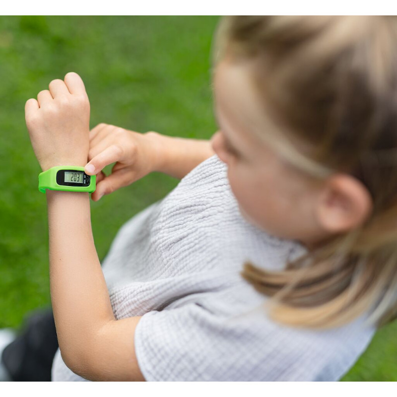 Ein Kind betrachtet im Freien auf dem Rasen eine grüne Digitaluhr an seinem Handgelenk.