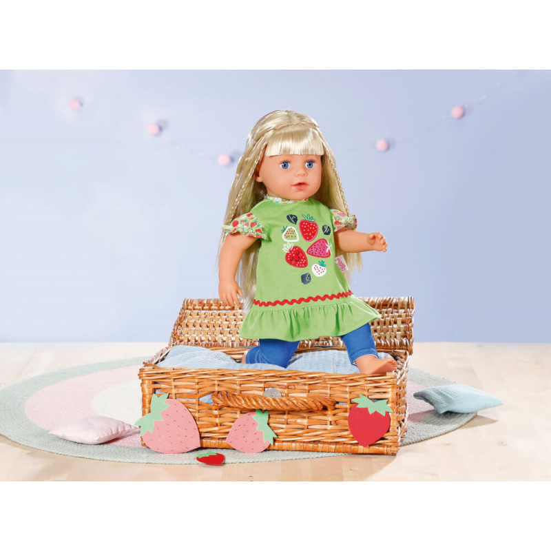 Eine blonde Puppe im grünen Erdbeerkleid sitzt in einem Korb auf einem pastellfarbenen Teppich.