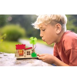 Junge bläst im Freien an einem sonnigen Tag auf ein kleines Holzhaus mit grünem Windrad.