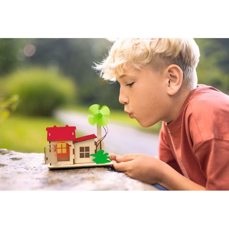 Junge bläst im Freien an einem sonnigen Tag auf ein kleines Holzhaus mit grünem Windrad.