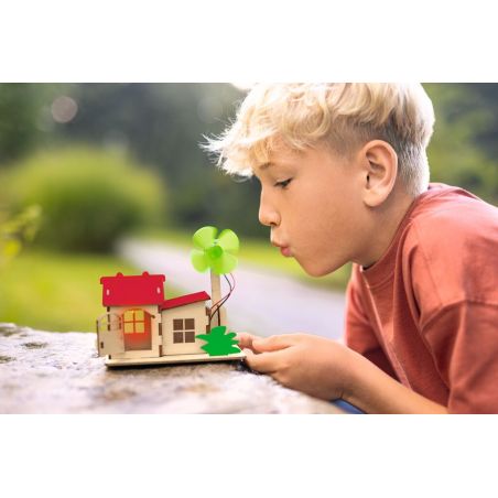 Junge bläst im Freien an einem sonnigen Tag auf ein kleines Holzhaus mit grünem Windrad.