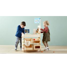 Zwei Kinder spielen mit einem Holzmarktstand auf hellem Holzboden vor einer hellblauen Wand.