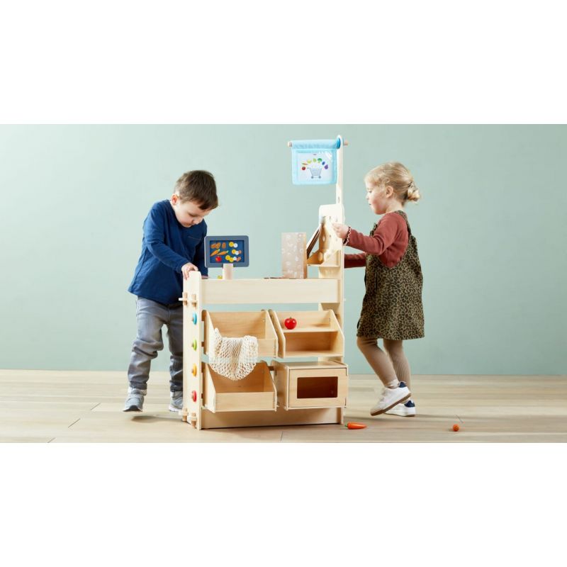 Zwei Kinder spielen mit einem Holzmarktstand auf hellem Holzboden vor einer hellblauen Wand.