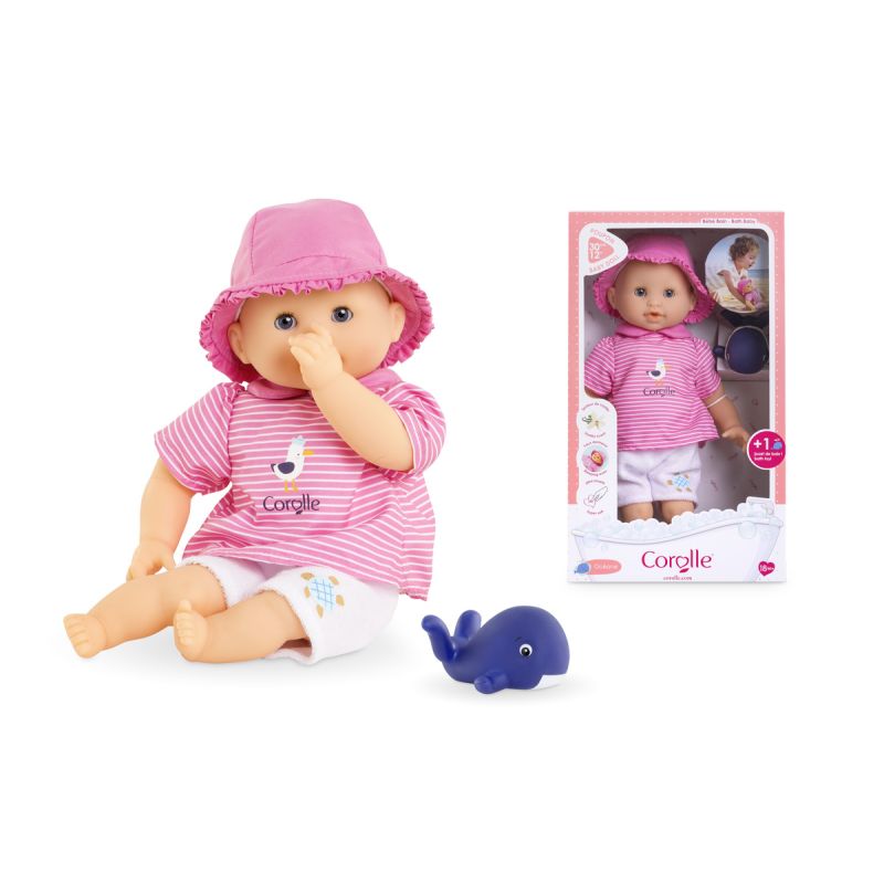 Eine Babypuppe in rosa Kleidung und Mütze sitzt mit einem blauen Wal-Spielzeug neben ihrer Verpackung. Eine Babypuppe in rosa Kleidung und Mütze sitzt mit einem blauen Wal-Spielzeug neben ihrer Verpackung.