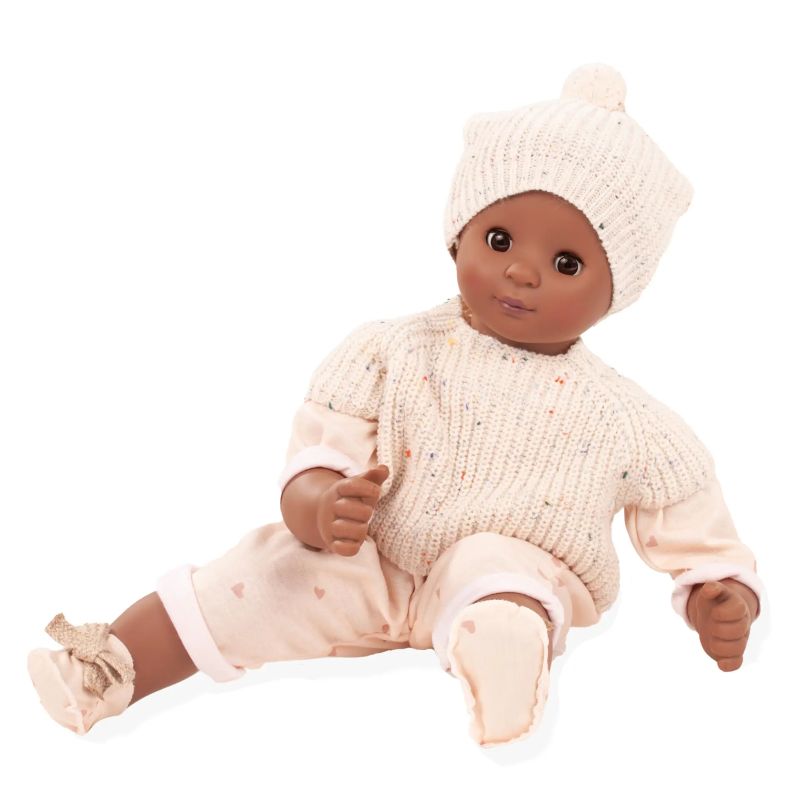 Eine braune Babypuppe mit cremefarbener Strickmütze, Pullover und rosa Hose sitzt aufrecht. Eine braune Babypuppe mit cremefarbener Strickmütze, Pullover und rosa Hose sitzt aufrecht.
