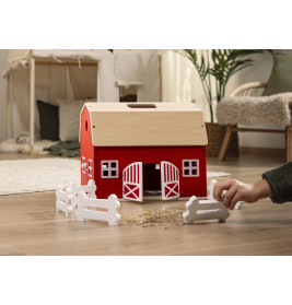 Eine Kinderhand stellt weiße Zäune um eine rote Spielzeugscheune auf einem Holzboden im Innenraum auf.