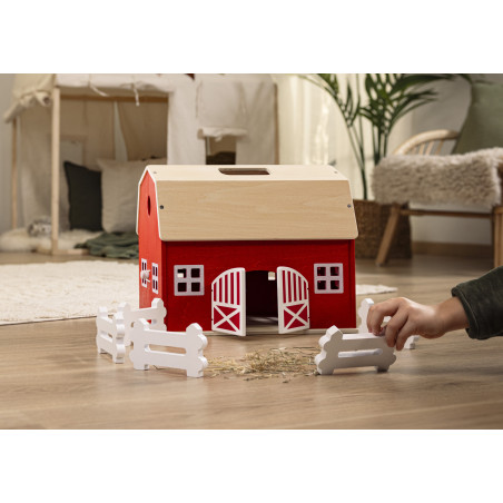 Eine Kinderhand stellt weiße Zäune um eine rote Spielzeugscheune auf einem Holzboden im Innenraum auf.