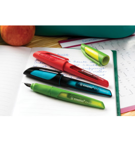 Drei bunte Füller liegen auf einem offenen Notizbuch, dahinter ein Apfel und ein Blatt Papier.