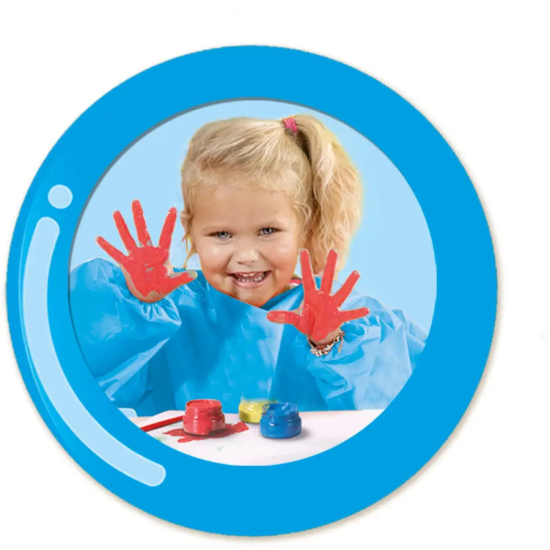 Kind im blauen Kittel lächelt, zeigt bemalte Hände, auf dem Tisch stehen Farbtöpfe.