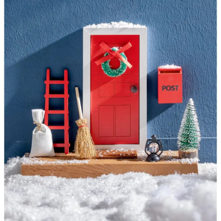 Miniaturrote Tür mit Kranz, Leiter, Besen, Briefkasten und Deko im Schnee vor blauer Wand.