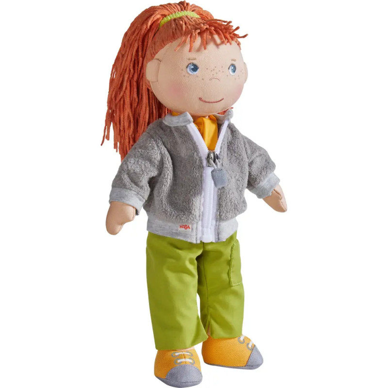 Stoffpuppe mit roten Garnhaaren, grauer Jacke, grüner Hose und gelben Schuhen steht aufrecht.