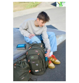 Eine junge Person sitzt auf Beton und greift nach einer Wasserflasche im grünen, mit Aufnähern verzierten Rucksack.