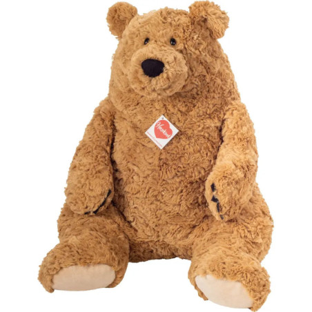 Großer brauner Teddybär mit lockigem Fell, beigen Pfoten und rotem Etikett sitzt aufrecht.