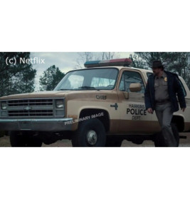 Ein Polizeichef steht neben einem beigen SUV der Hawkins-Polizei, im Hintergrund Bäume.