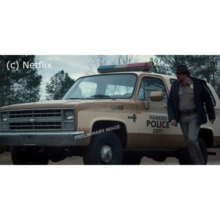 Ein Polizeichef steht neben einem beigen SUV der Hawkins-Polizei, im Hintergrund Bäume.