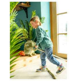Lächelndes Kind reitet drinnen auf einem Plüsch-Dinosaurier bei Pflanzen am sonnigen Fenster.