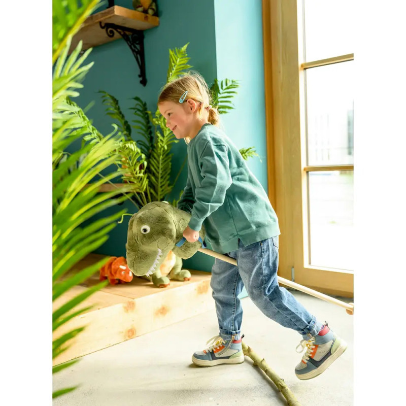 Lächelndes Kind reitet drinnen auf einem Plüsch-Dinosaurier bei Pflanzen am sonnigen Fenster.