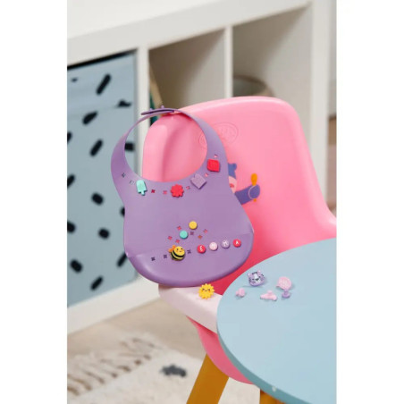 Ein lila Lätzchen mit bunten Anhängern hängt an einem rosa Hochstuhl neben einem blauen Tisch im Spielzimmer.